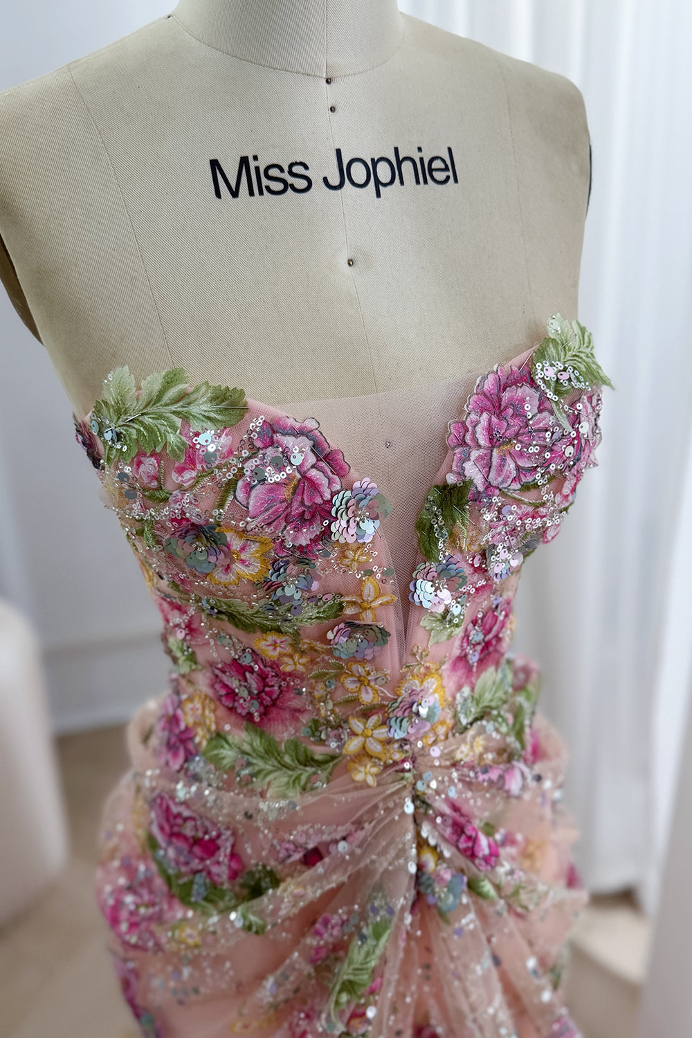 Le gros plan du buste dévoile des fleurs fuchsia, un feuillage vert, des paillettes pastel et un empiècement de pudeur sous un décolleté plongeant en cœur, témoignant d’un savoir-faire artistique.