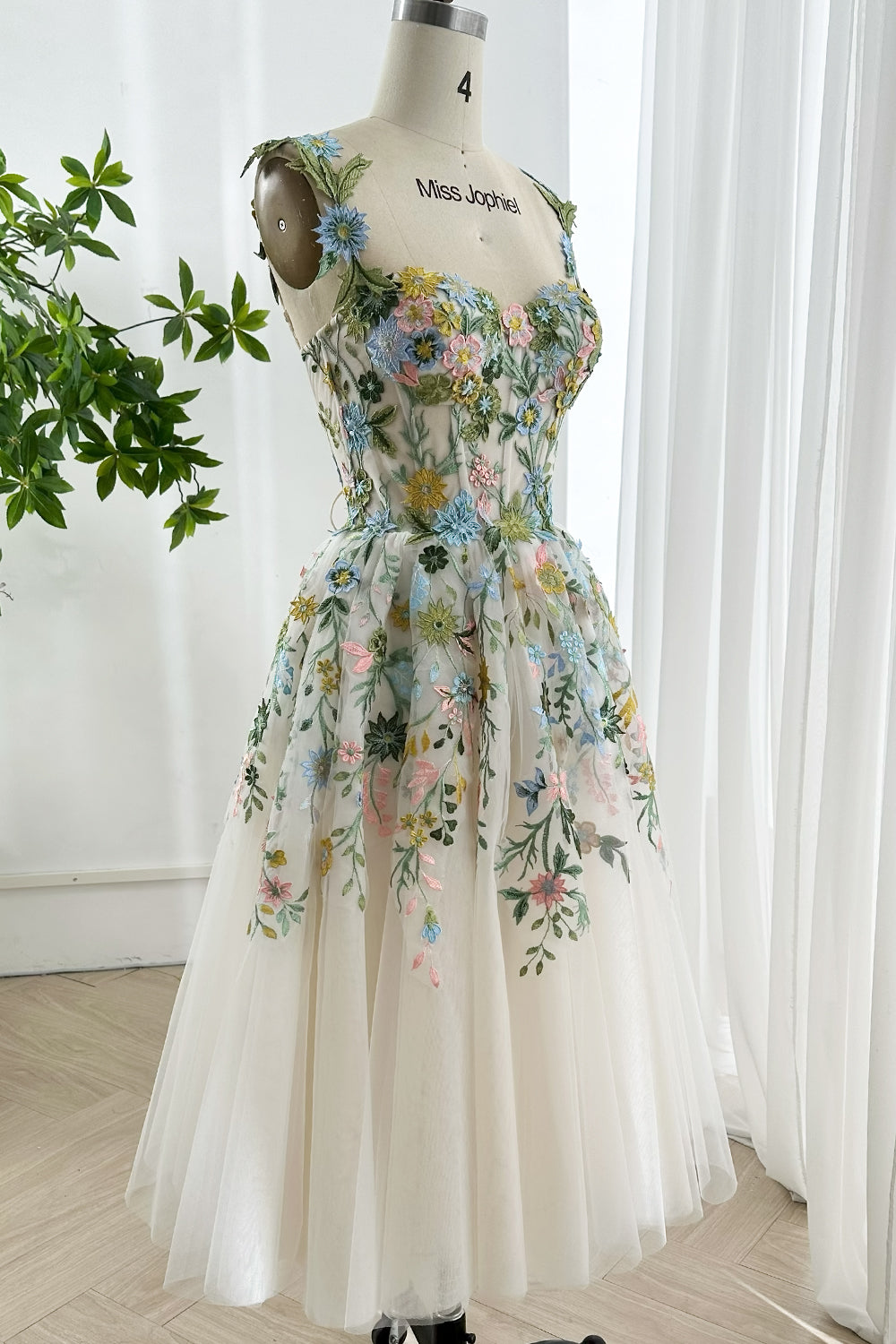 Elegant pastel midi dress embroidered with delicate tulle layers soft botanical motifs displayed elegantly on mannequin by window