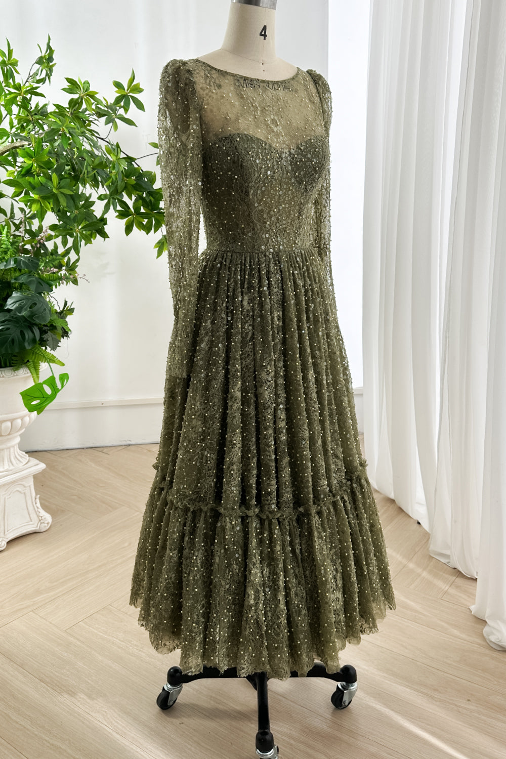 Elegant olive lace midi gown featuring illusion neckline long sleeves fitted waist and A-line skirt shown on dress form in bright studio