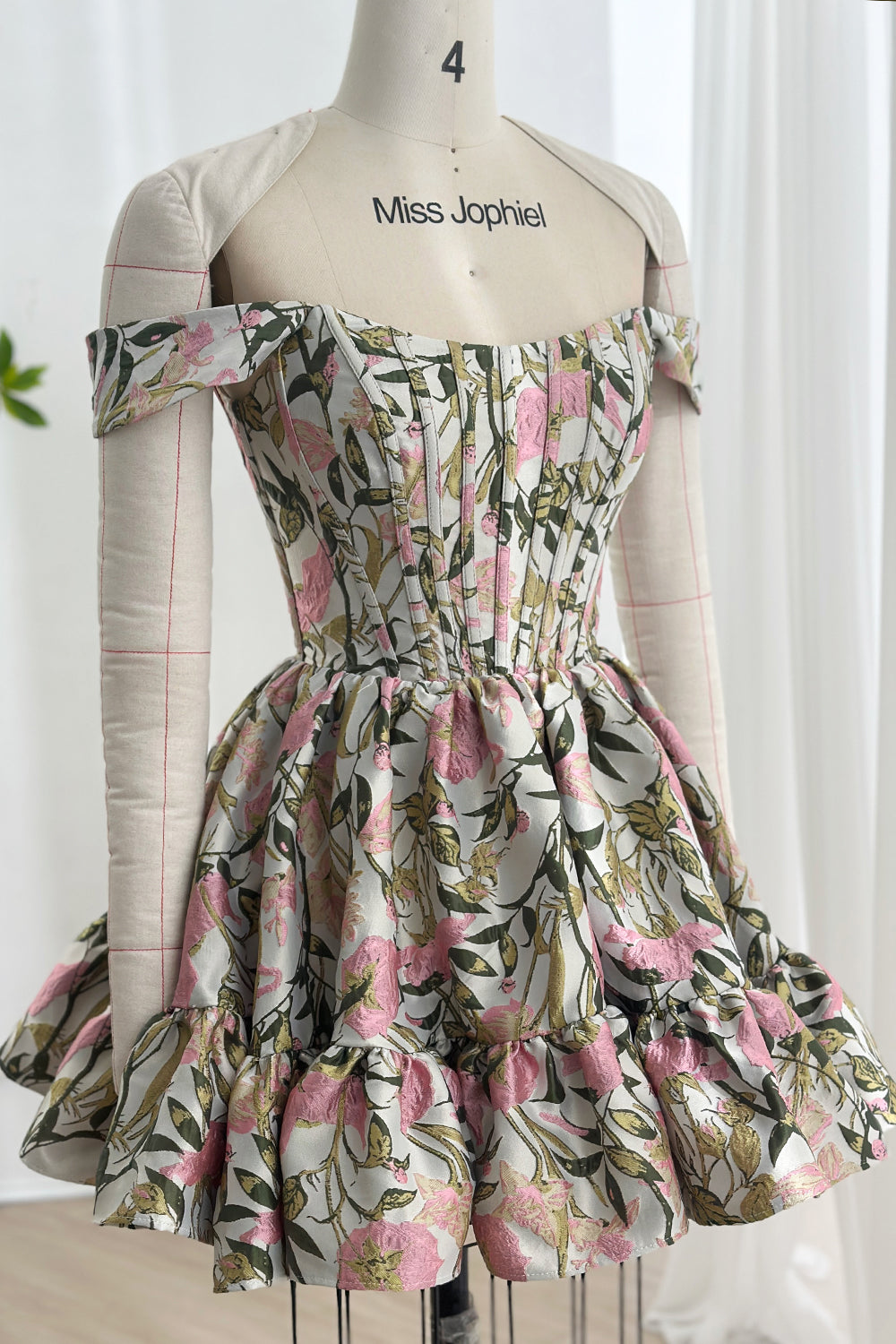 Structured bodice mini featuring blush blossoms sage foliage jacquard texture sculpted waistline ruffled hemline couture presentation mannequin studio