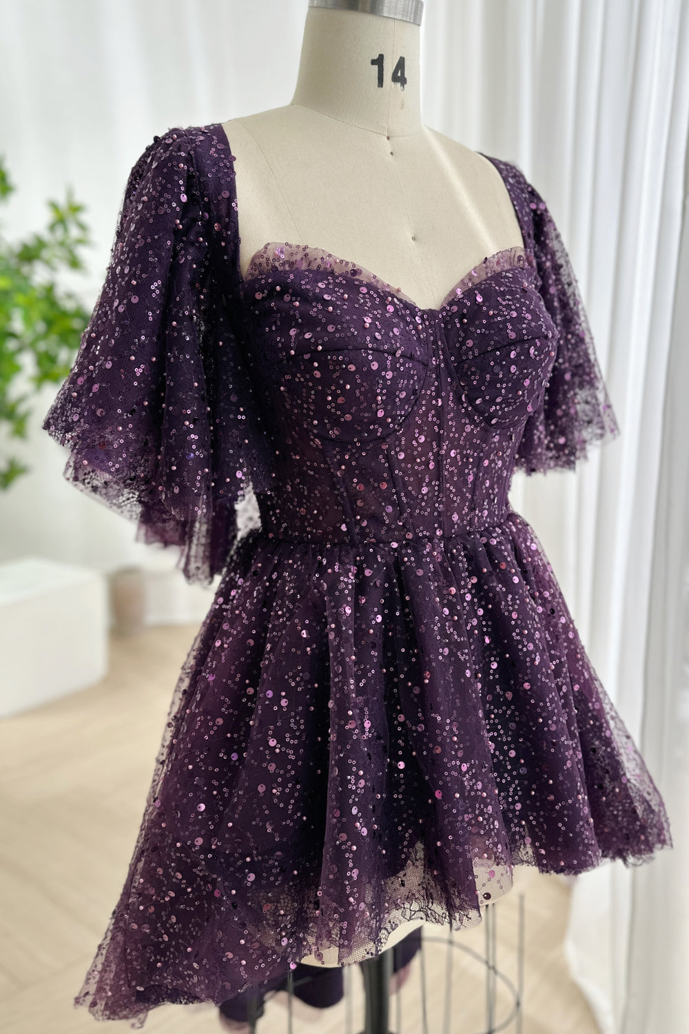Plum glitter tulle corset mini dress displayed mannequin structured bodice flutter sleeves high low skirt sparkle texture studio setting