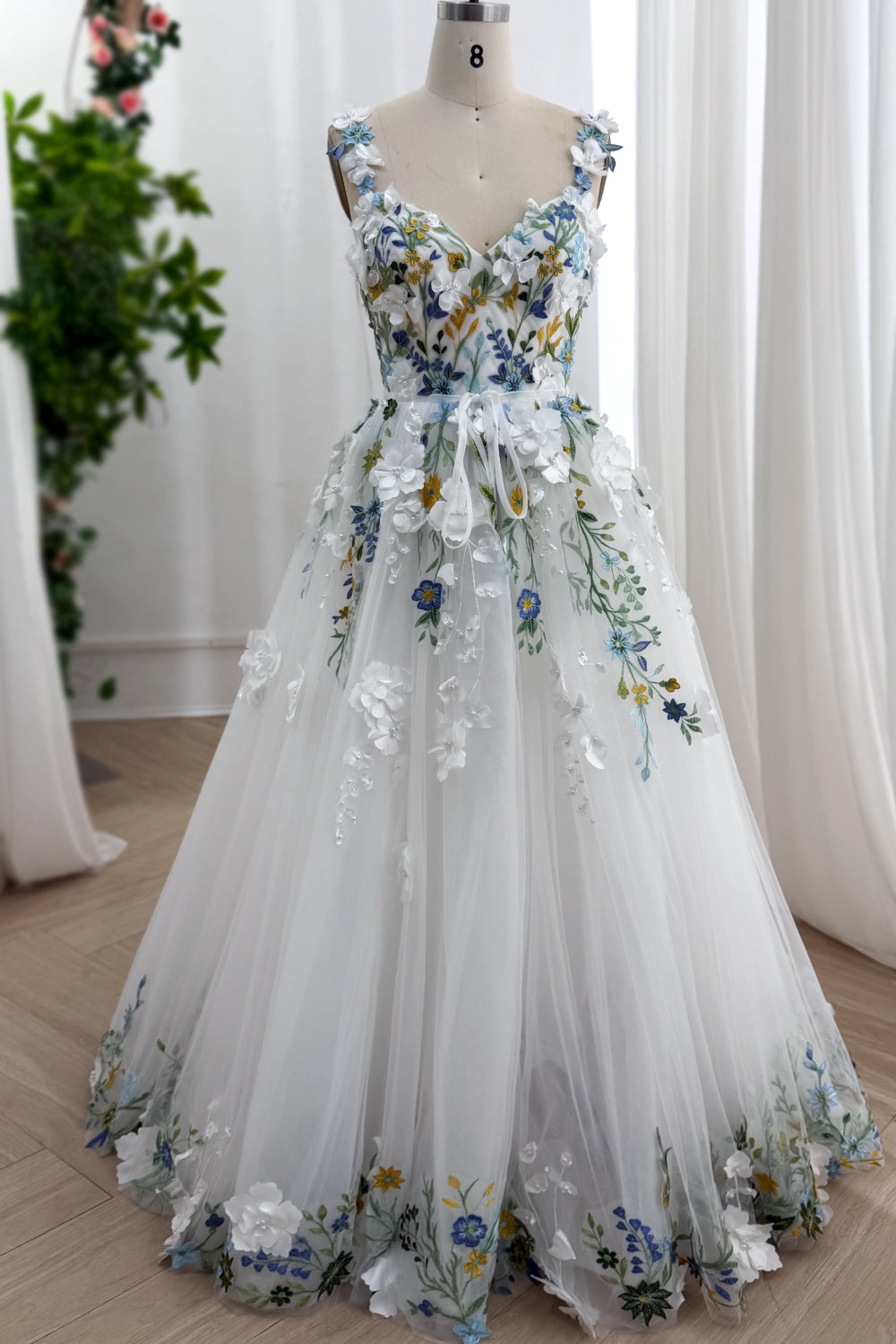 Ethereal ivory gown showcasing dimensional blossoms azure embroidery meadow motifs graceful silhouette arranged upon atelier mannequin