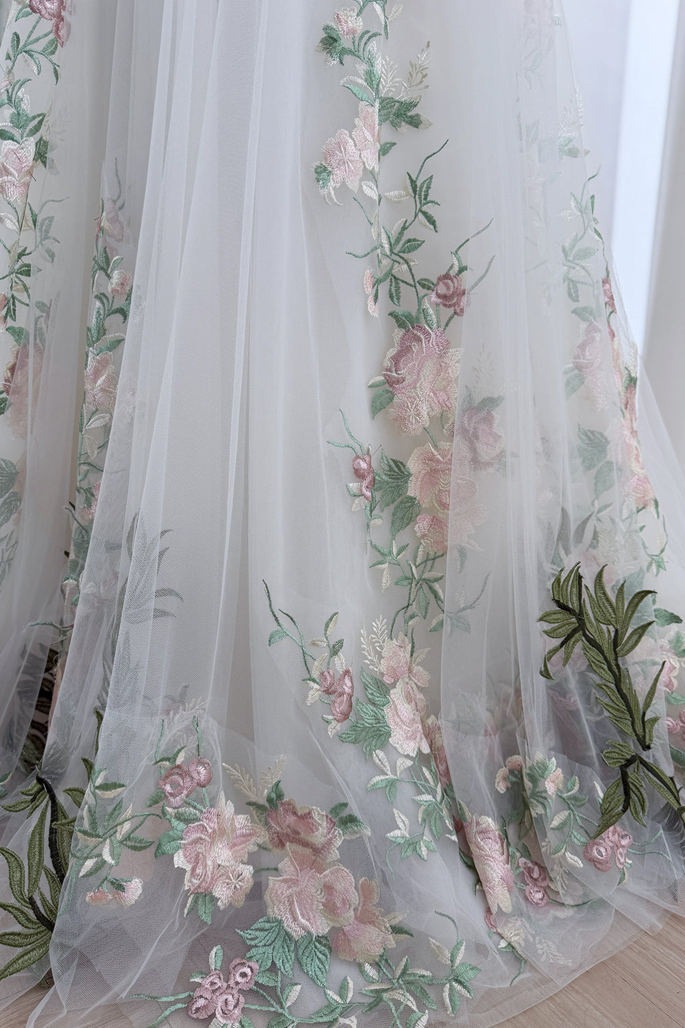 Hemline detail displays tiered mesh, wildflower motifs, rising stems, soft petals, translucent overlay, floor-length texture.