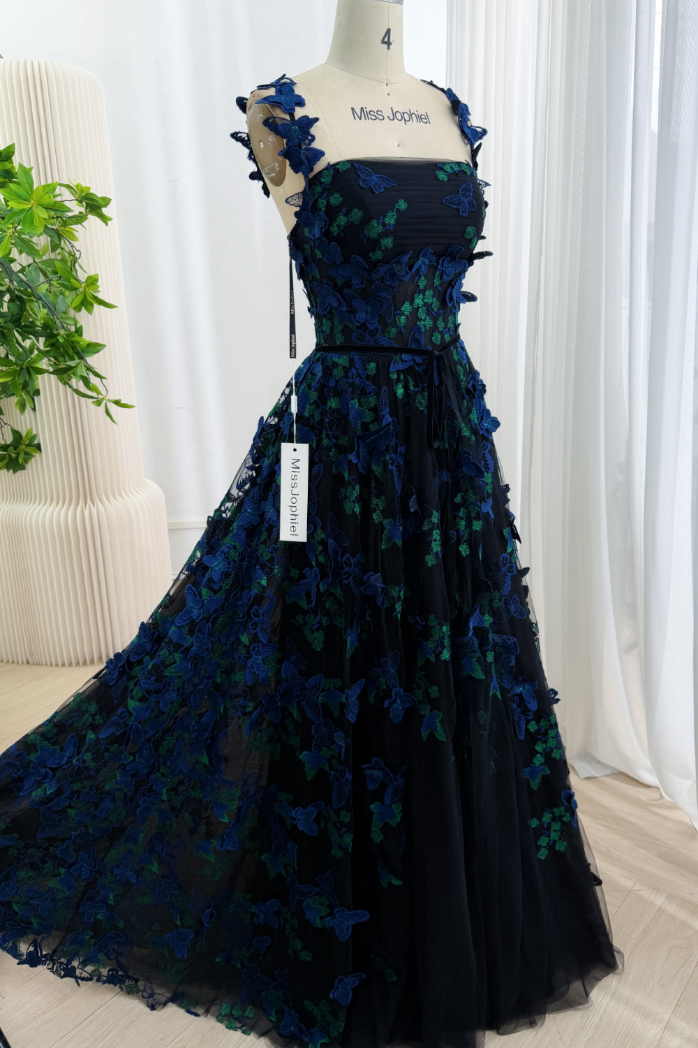 Front view of a black floor-length A-line gown with blue and green 3D butterfly appliqués and a ribbon waist tie