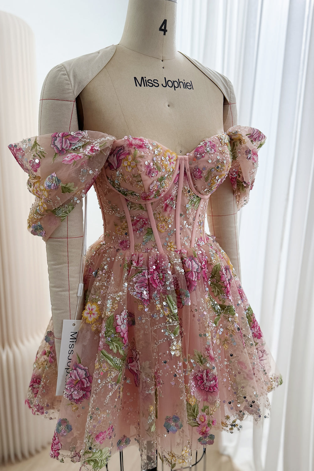 Angled front view of a blush pink embroidered tulle mini dress showing draped off-the-shoulder sleeves and a fitted corset bodice.
