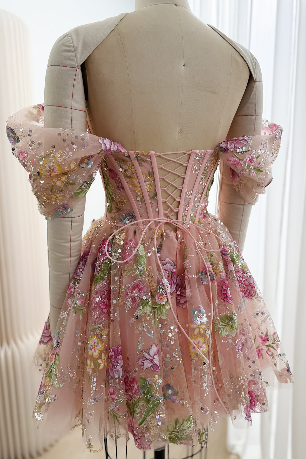 Back view of a blush pink floral sequin mini dress with pink corset lacing, off-the-shoulder sleeves and a gathered flared skirt.