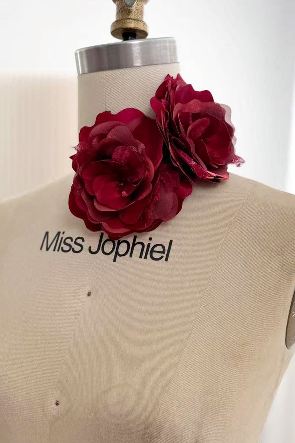 Two burgundy fabric rose flowers with layered sheer and lustrous petals displayed at the neck of a dress form.