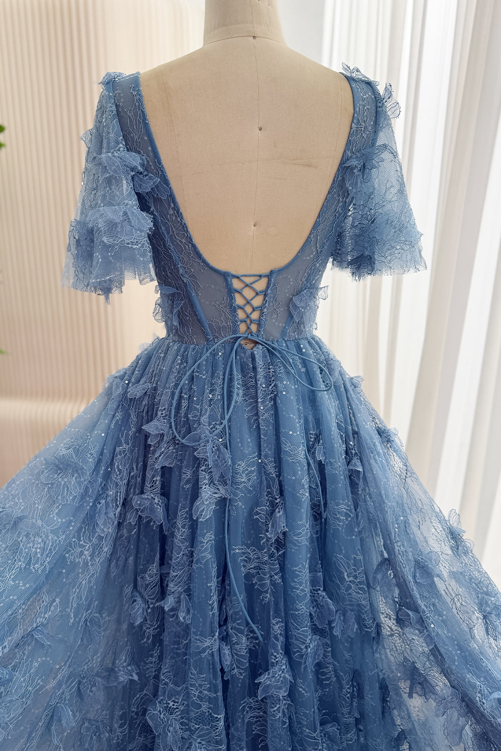 Back detail of dusty blue floral lace gown showing a low scoop back, lace-up fastening, sheer sleeves, and raised floral appliqués.