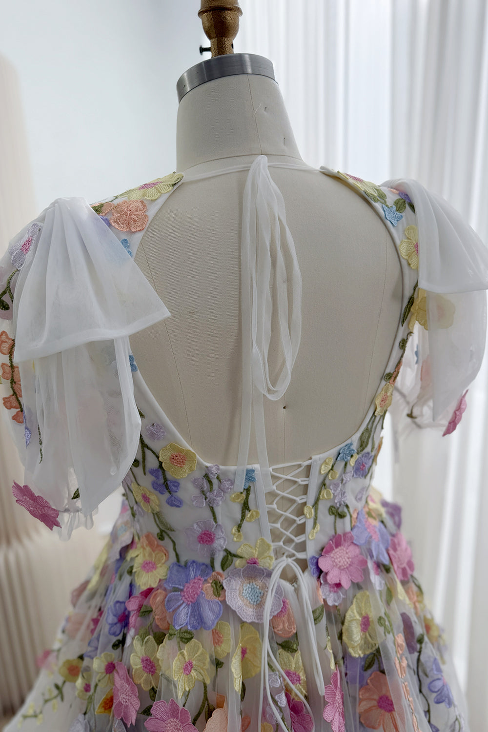 Close-up of the floral wedding dress back showing corset lacing, a low open neckline, an upper-back tie, and sheer puff sleeves.