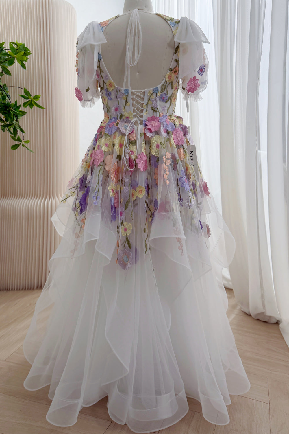 Back view of a white floral wedding dress with a low open back, corset lacing, sheer puff sleeves, and a floor-length ruffled skirt.