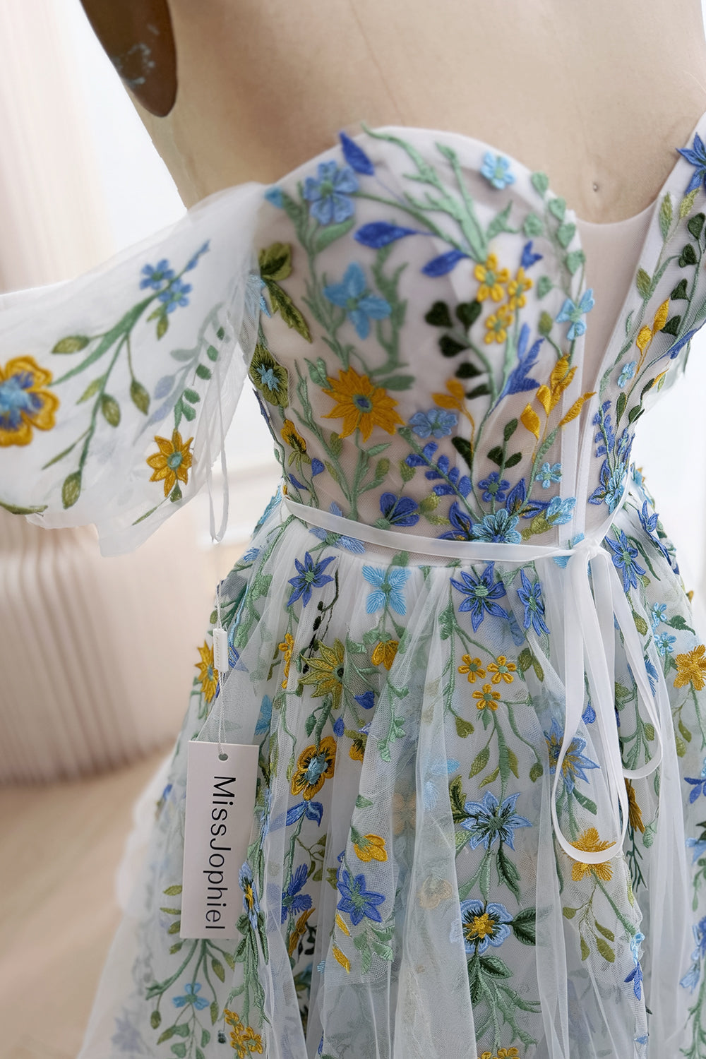 Close-up of blue and yellow wildflower embroidery with green leaves across the ivory gown's bodice, sleeve, and ribbon-tied waist.