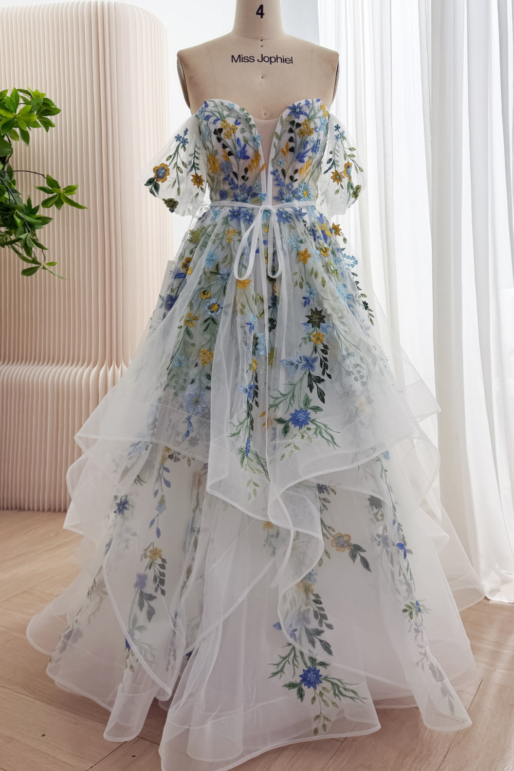 Front view of an ivory wildflower embroidered tulle gown with off-the-shoulder flutter sleeves, ribbon waist tie, and cascading skirt ruffles.