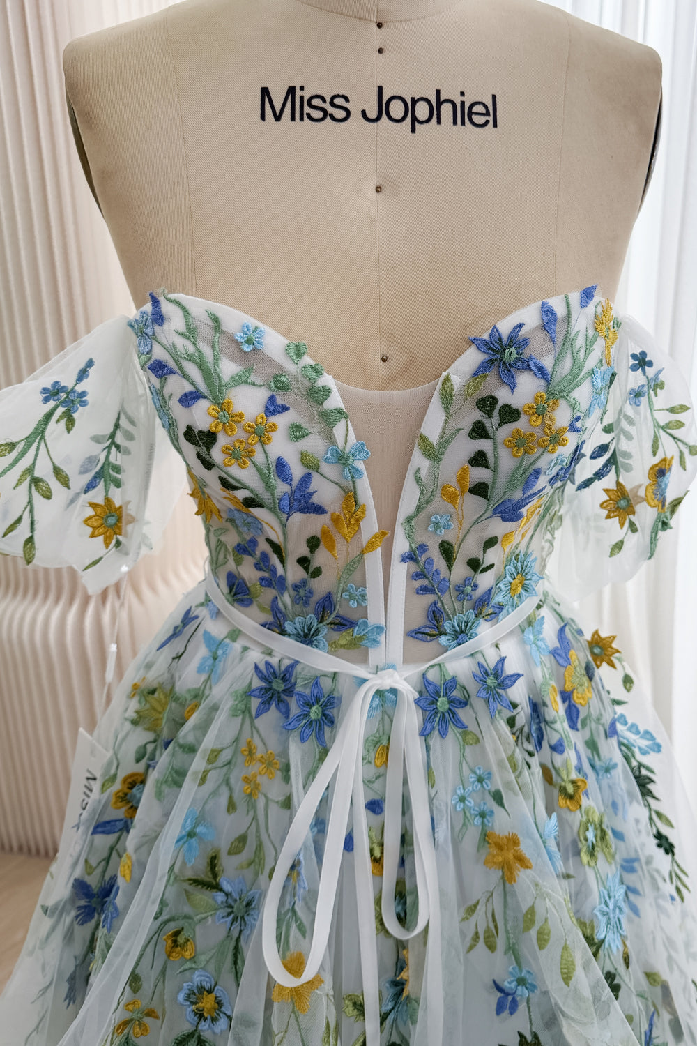 Front bodice detail of the ivory wildflower gown with a sweetheart neckline, deep illusion plunge, blue and yellow embroidery, and ribbon bow.