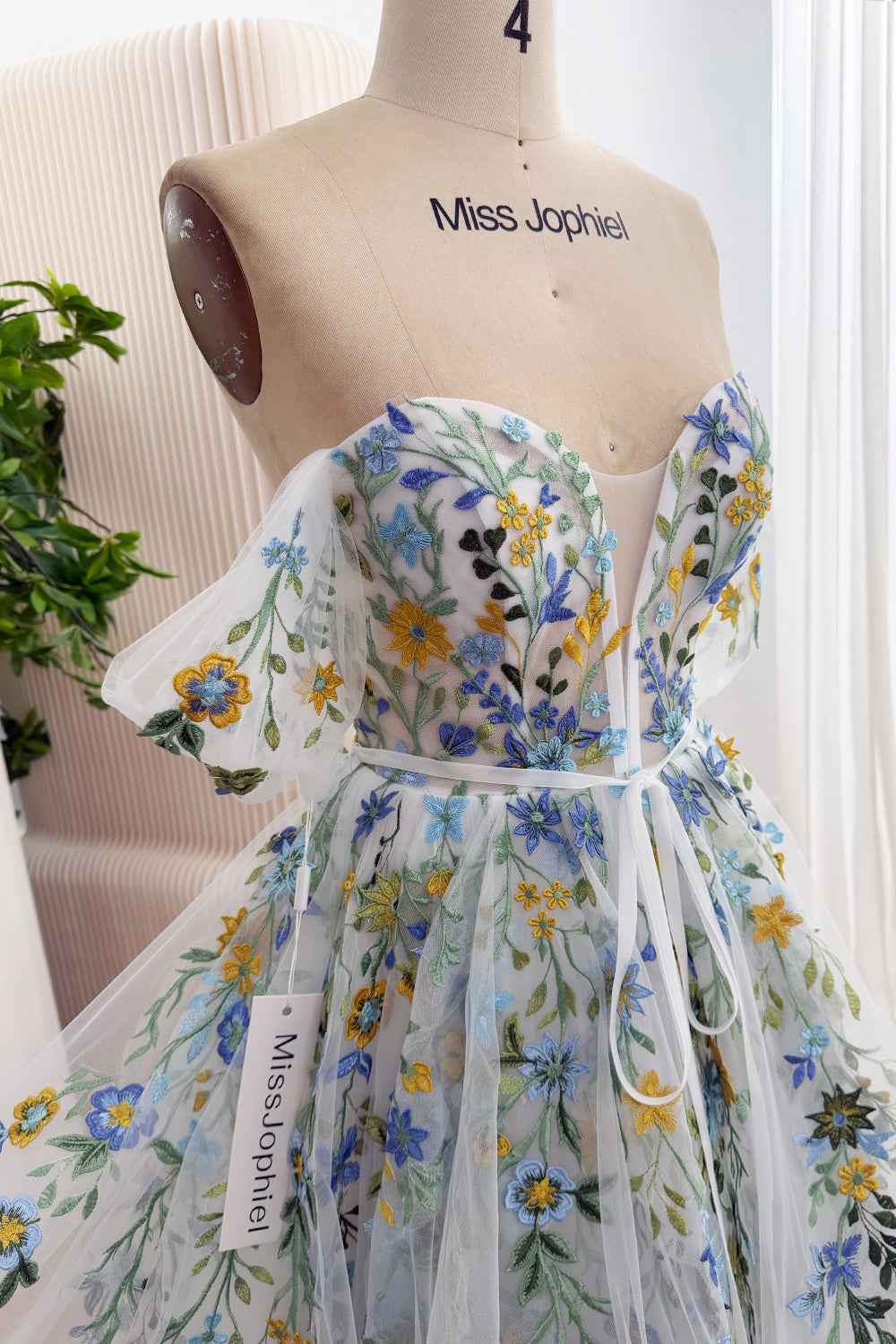 Angled close-up of the ivory floral gown showing blue and yellow embroidery, an illusion plunge neckline, and an off-the-shoulder flutter sleeve.