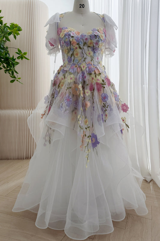 Front view of a white wedding dress with pastel floral embroidery, a sweetheart bodice, sheer puff sleeves, shoulder bows, and cascading skirt ruffles.