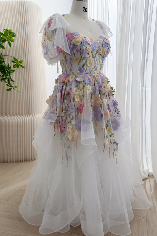 Angled view of a pastel floral wedding dress showing dimensional flower appliqués, shoulder bows, and a full skirt with cascading sheer ruffles.