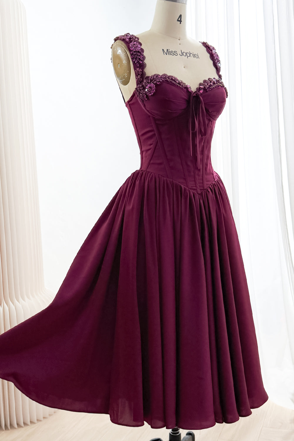 Angled front view of plum tea-length A-line dress with embellished straps, sweetheart corset bodice, and full skirt.