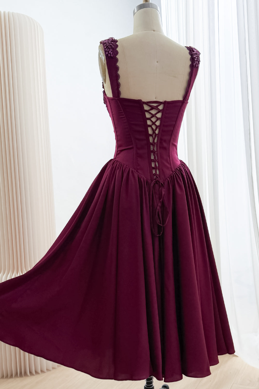 Back view of plum tea-length A-line dress with embellished shoulder straps, lace-up corset, and full gathered skirt.