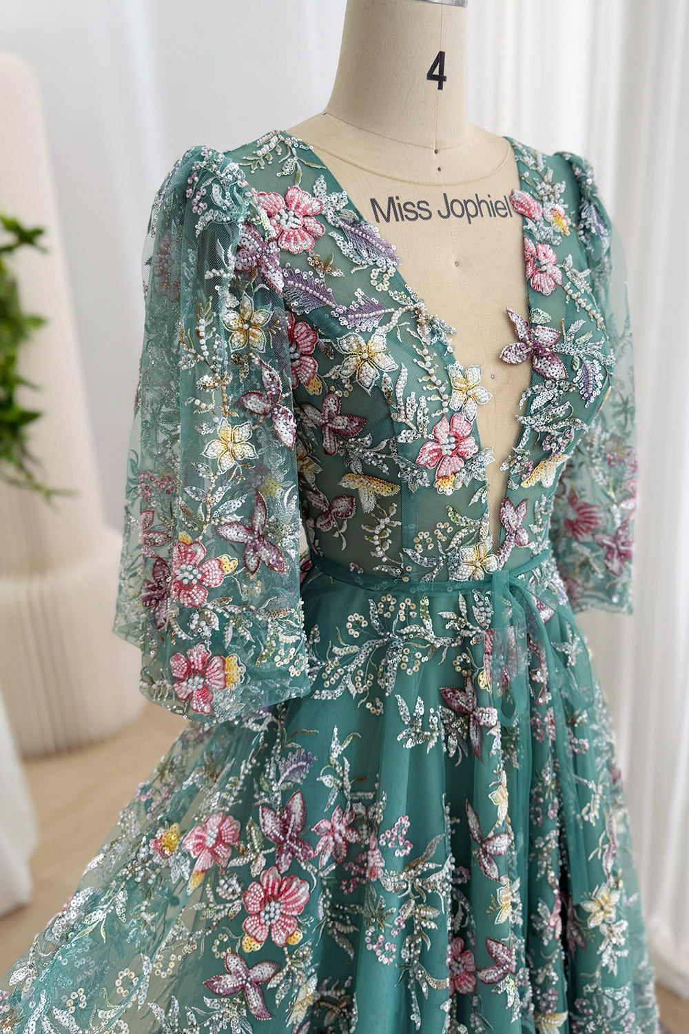 Close-up front view of a teal tulle gown with a plunging illusion V-neckline, multicolor floral embroidery, sequins, and sheer sleeves